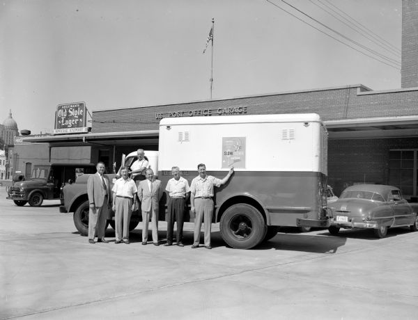 A new red, white and blue mail truck is displayed at the Madison Post Office garage. A poster on the side of the truck reads: "Don't be a Hurry Bug — Slow Down and Live." The poster is part of a nationwide "Slow Down and Live" traffic safety campaign. Standing in front of the truck are (left to right): Asst. Postmaster LeRoy Murray; post office garage superintendent Leslie Doyle; mechanic Fred Grant; John Whitmore, safety director; Henry Olson, dispatcher and superintendent of driver safety; and Don Cowen, clerk of vehicle services.      