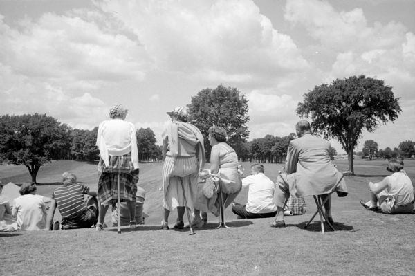 A group of men and women watching the golf match at the 26th Annual Women's Western Open golf championship at the Maple Bluff Country Club. A few people are sitting on four different types of small portable chairs.