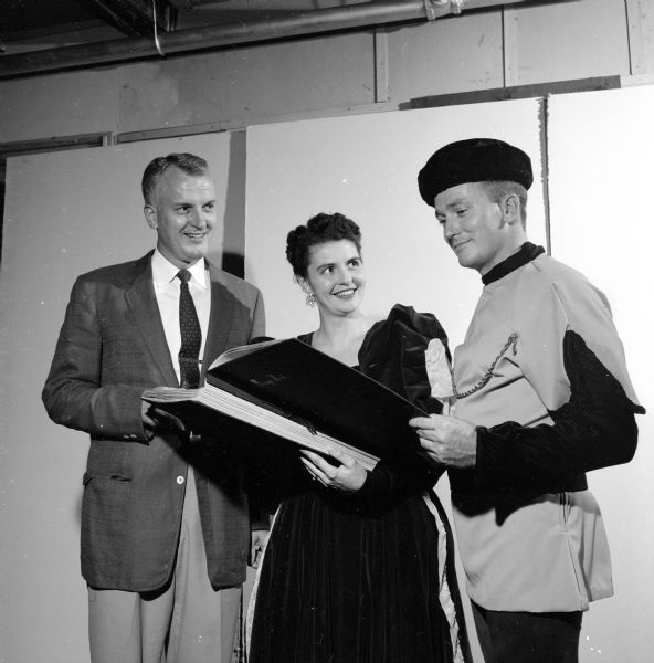 Joseph Klein, Lois Klein and Marvin Foster (left to right) are examining a scrapbook depicting the first ten years of activity for the Madison Theatre Guild. Mrs. Klein and Mr. Foster are shown in costume.