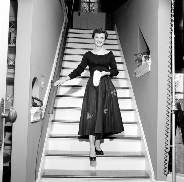 Clothing outfits available at Madison stores being modeled by young women:  Standing on a flight of steps, Margaret McDowell is wearing a black felt, full circle skirt with strawberry appliques and a black, off-the-shoulder bulky, ribbed knit sweater. She is holding a pair of white gloves. 