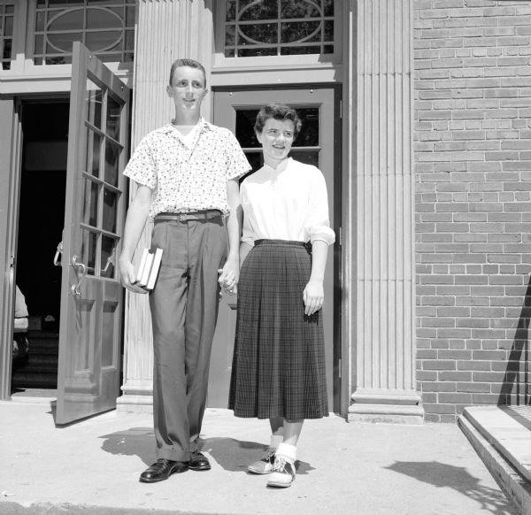 Clothing outfits available at Madison stores being modeled by young women: Margaret McDowell is wearing a pleated wool and orlon skirt in Black Watch Tartan, a worsted jersey blouse with a mandarin collar, and saddle shoes. Kent Jones is holding her hand. They both attend West High School.