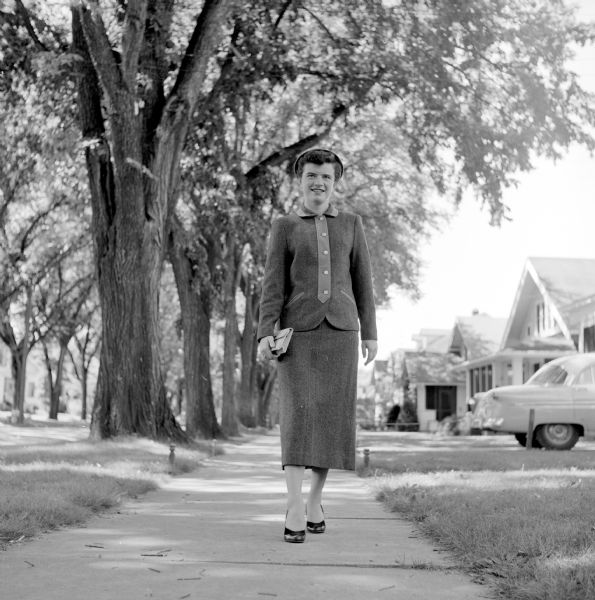Clothing outfits available at Madison stores being modeled by young women: Walking on a sidewalk, Margaret McDowell is wearing a suit of a avocado green tweed with solid panels of the same color. The box jacket buttons form a Peter Pan color. She is also wearing high heeled shoes and a "pert" little hat. Walking on a sidewalk in a residential neighborhood, she is carrying a pocketbook.
