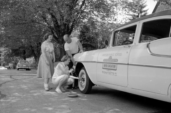 West High School driver education instructor and industrial arts teacher Dalton Hedlund (of 114 Leon Street), is teaching Judy Davis, (left, of 2001 Van Hise Avenue) and Kathy Kingsburyl (kneeling, of 575 Toepfer Avenue) how to change a car tire at the side of a road. The sign on the car door reads, "Madison Public Schools, Driver Education, Pyramid Motor Co."