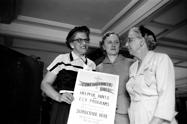 Three women are holding a poster at a convocation session of the Wisconsin Congress of the Confraternity of Christian Doctrine at St. Raphael Cathedral. The poster reads: Confraternity Call - Helpful Hints on CCD Programs [Confraternity of Christian Doctrine] - Subscribe Here.