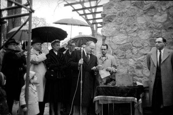 Participants attending in a cornerstone laying ceremony for the B'nai B'rith Hillel Foundation Building located at University of Wisconsin on a rainy day. They held the ceremony even though the cornerstone had been stolen by vandals. Standing (left to right) are: Ben Glass; Mrs Louis Behr of Chicago; Lewis Phillips of Eau Claire; Ben Rosenberg of Green Bay; Gordon Sinykin; and Rabbi Max Ticktin.