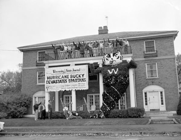 A neo-classical styled fraternity house on Langdon Street. Fraternity brothers, waving, fill an upper balcony above four large pillars, from which a sign is suspended that looks like the Wisconsin State Journal front page with a headline that reads: "Hurricane Bucky Devastates Spartans." Beneath the headline are words about: "City orders Langdon Street Slum Cleanup." Beside it is a large, two-story Bucky Badger made of paper emerging out of a funnel.
