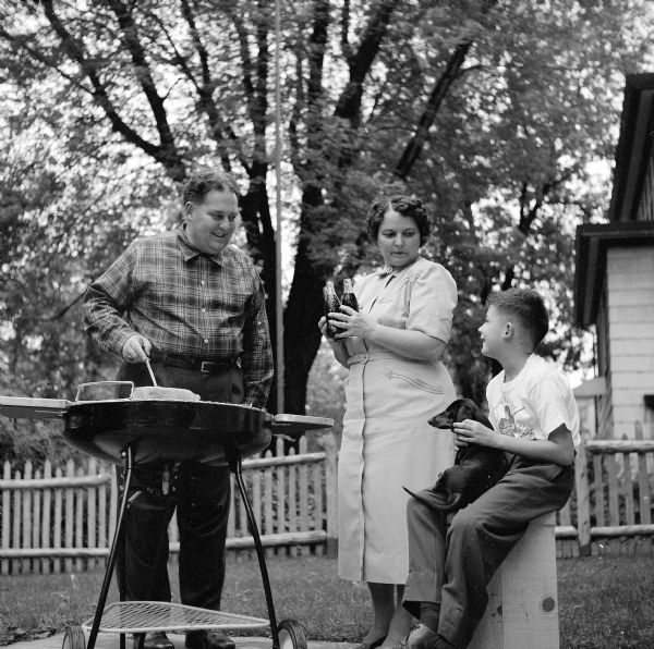 A mother and son look on as the father tends to a thick steak cooking on a portable barbecue. The boy is holding a Dachshund puppy; the mother, three bottles of beer. They are on a concrete patio in a backyard, enclosed by a picket fence. A tree in the background is standing in the neighbor's yard. 