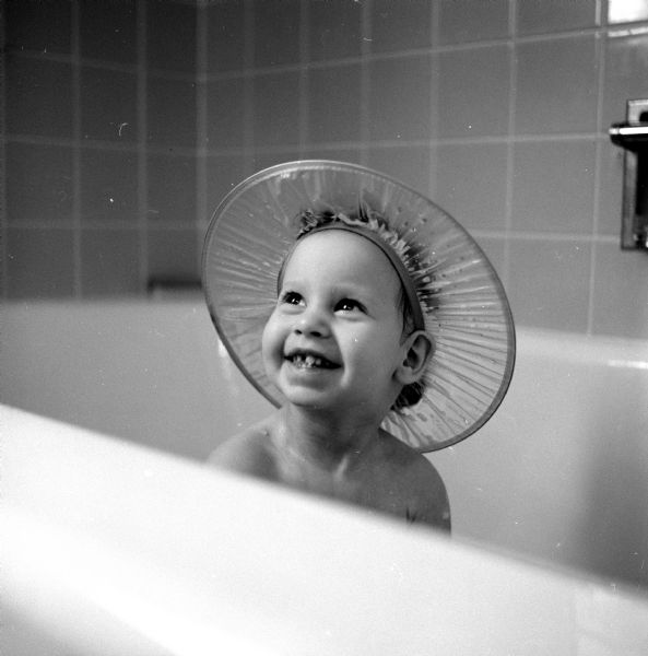 The photographer's daughter, Laurie Stein, posing in a bath tub wearing a hat consisting only of a wide rim encircling her head to prevent shampoo from getting into her eyes.