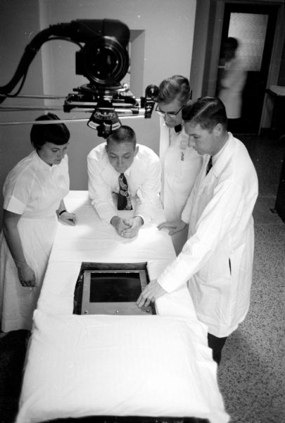 Slightly elevated view of medical staff examining an angio-cardiograph X-ray camera are (from left): Yvonne Traeder, an X-ray technician; Dr. I.A. Krystosek, resident in radiology; John H. Juhl, Chief of Radiological Services at the VA Hospital; and Dr. Charles Crumpton, Director of the University Cardiovascular Lab.