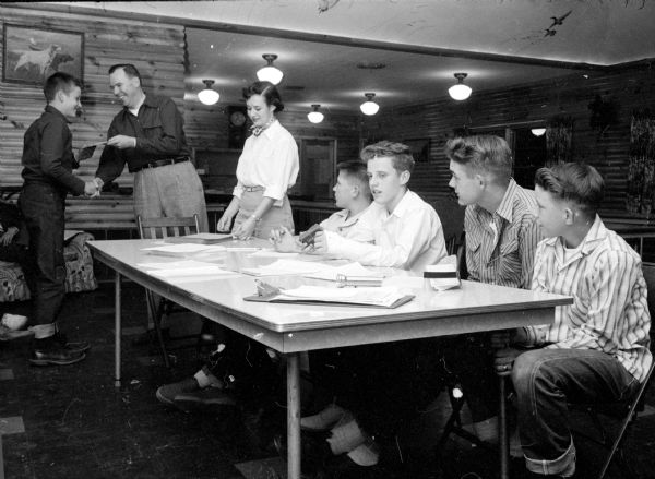 Club officers in a meeting. At left, Roger Bell is receiving an award from the National Rifle Association (NRA) presented by M.D. Doyle, instructor at the University of Wisconsin and instructor for the Junior Rifle Club. Standing in the middle is Hallie Hohf, Club Secretary. Sitting at a table are: Jack Hobi, vice president; Jon Gruber, president; and Gene Stormer, executive officer.