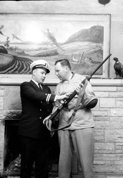 Lt. William Ruddick, Commander of the Navy and Marine Corps Training Center in Madison, checks a gun with instructor M.D. (Pat) Doyle. They are standing in front of the Club's fireplace that has a stuffed pheasant on the mantle, and a large painting of these birds in flight in a rural landscape.