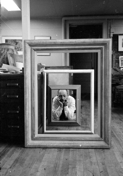 University of Wisconsin artist-in-residence, Aaron Bohrod, posing in his studio behind three vertically nested picture frames.