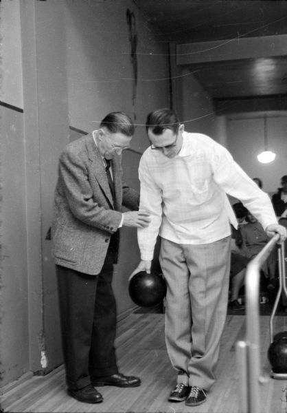 Jim Sletten (in dark glasses) is holding onto a guide rail with his left hand while holding a bowling ball in hi right hand as he accepts assistance from Lions Club member, Oscar Jensen. Jim is one of about fifteen visually impaired participants who gather weekly on Tuesdays at Playdium Alleys located at 112 North Fairchild Street. None of the participants has vision better than 10 percent.