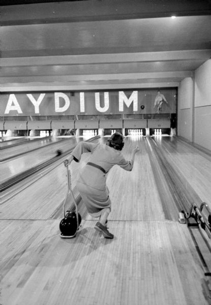 Hazel Buege is holding onto a rail with her left hand, and releasing a bowling ball with her right hand towards standing bowling pins. She is one of about fifteen visually impaired participants who gather weekly on Tuesdays at Playdium Alleys located at 112 North Fairchild Street. None of the participants has vision better than 10 percent.