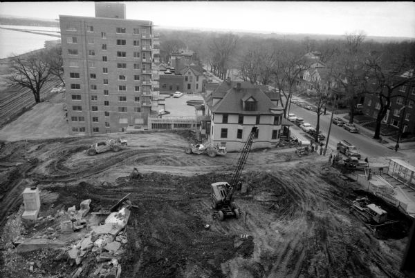 Elevated view from the top of central section of the State Office Building at 1 West Wilson Street, showing earth moving equipment at the excavation site for the new west wing of the State Office Building. The new multi-story building is Town House Apartments at 111 West Wilson Street, and the house in the center foreground will be demolished.