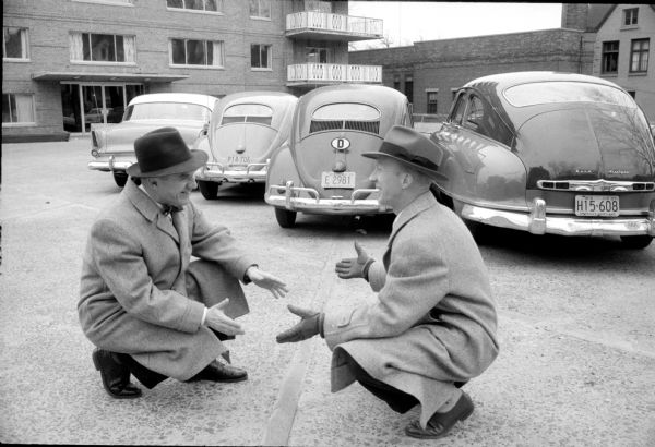 Two men crouching in a parking lot smiling while arguing over who gets to park in one parking spot, with their respective cars fender to fender behind them. One of them drives a Volkswagon Beetle.