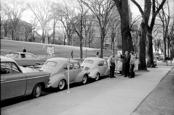 Two drivers are flipping a coin to determine who feeds the meter with their two small cars squeezed into one parking stall on North Park Street by Bascom Hill at the University of Wisconsin. The cars appear to be Renault 4CVs. The building in the distance (center) is Science Hall.