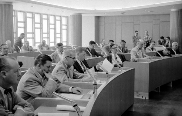 The Madison City Council holds its reorganization meeting in the new City-County Building Council Chamber. Shown seated from left to right are: in the first row, Forsythe, Perkins, McCormick, Fitzpatrick, and Elder; and in the second row, Hagan, Henry, Hernan, Nuckles, and Spohn. In the rear row are Water Superintendent Elmer L. Nordness (left), Building Superintendent Ray F. Burt (third from left), and Mayor Assistant Hugh D. Ingersoll (far right).