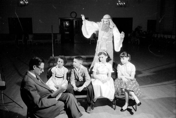 The Madison Women's Club presents the original play "Man and His God" to raise money for scholarships. A group from Beth El Temple rehearsing a scene in which a teacher in the synagogue is reading to pupils, as Isaiah, one of the Old Testament prophets, appears behind them in the classroom. The "teacher," Laurence A. Weinstein of 1134 Waban Way, is sitting with four child actors (left to right) of the theatrical performance: Susan Stone of 645 Odell Street; Dan Weinstein of 1134 Waban Way; Lisbeth Kline of 4214 Cherokee Drive; and Susan Shapiro of 3710 Odana Road. "Isaiah" is played by Ralph E. Kline, Lisbeth's father.
