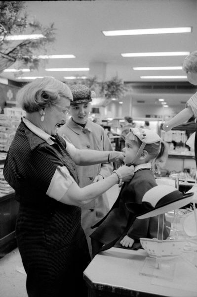 Jo Clark, age 3, is sitting on a table in a department store as clerk Frances Sternberger (left) is tying the chin strap of a new Easter hat. Jo's mother, Janis Clarke, also wearing a hat, is smiling approvingly.