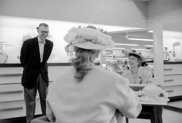 Arthur Dier smiles approval as his fiancee, Evelyn Schaefer, asks his opinion of a bright flowery Easter hat at a Madison store. Ms. Schaefer, in the foreground, is seen facing away, wearing a hat and sitting at a mirror that is reflecting her expression as she is exchanging glances with her fiance.