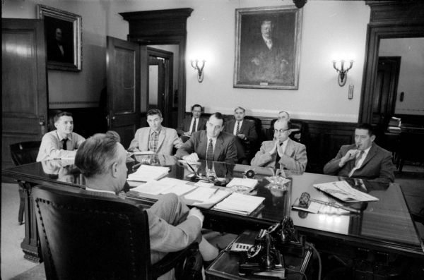 Wisconsin Governor Vernon W. Thomson sits at his desk during a press conference. Newsmen seated around the governor's desk are (from the left): Mack Hoffman, <i>Associated Press</i>; Richard Bradee, <i>United Press</i>; Robert E. Doyle, <i>Milwaukee Journal</i>; Richard A. Brautigam, <i>Milwaukee Sentinel</i>; and Lew Roberts, <i>Wisconsin State Journal</i>. John Wyngaard, <i>Green Bay Press Gazette</i>, attended but was out of the camera view. Seated in back along the wall are three members of the governor's staff: Philip Sellinger, researcher; W. Donald Knight, financial advisor; and former Assemblyman Alfred Ludvigsen, liaison with the legislature.
