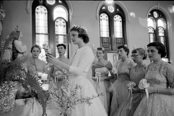 Veronica Goebel (of Racine) is carrying the crown of May flowers to place on the head of a Virgin Mary statue. Maids of Honor in dresses holding posies are (left to right): Mary Goodman (of Antigo); Carole Mueller (of Sheboygan); Mary Ellen Buedel (of Chicago); Ruth Bosch (of Appleton); Barbara Belds (of Sun Prairie); and Mary McCurville (of Darlington). A fern plant and freshly cut branches from a blooming apple tree stand in a vase by the altar.