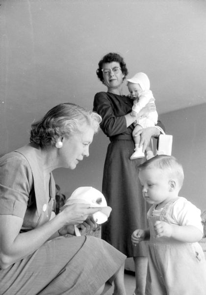 Mary Gill, a member of the Madison Advisory Board of the Children's Service Society, a Red Feather agency, is about to put a hat on one of the three foster children being cared for in the home of Mr. and Mrs. Edward Check, Route 1. Mrs. Check is holding one of the two foster infants currently sharing her home.
