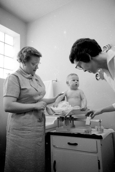 Foster mothers take homeless infants and children into their homes. Members of the Madison advisory board of the Children's Service Society, a Red Feather agency, drive the foster mothers and their babies to a clinic for medical checkups one morning each month. Ricky's foster mother, Mrs. Haugen, supports him on the scales as he looks at Nurse Esther Wardale who is adjusting the weight.