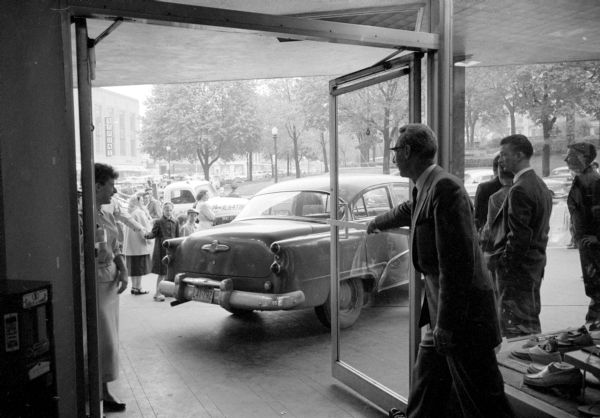 View from inside store of Ray Greene, shoe department manager for Karsten's men's store on Carroll Street on Madison's Capitol Square, standing in the doorway and looking at a car that had rolled across the street driverless after being parked. Three men had stopped the car before it reached the storefront and plate glass windows. The trees surrounding the Wisconsin State Capitol are in the background.