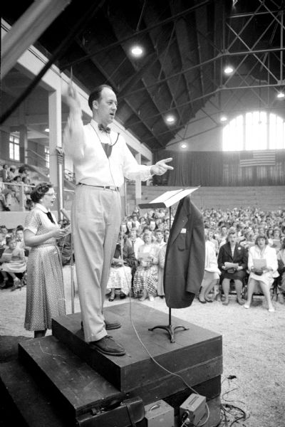 Warren Wooldridge conducts the 24th Annual Radio Music Festival of the WHA Wisconsin School of the Air concert held at the U.W. Stock Pavilion attended by 2,019 students. Behind him at the microphone is soloist Mrs. Warren Wooldridge from his "Let's Sing" broadcasts. The concert was broadcast live over WHA Radio.