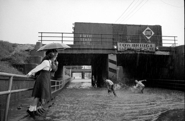 Children frolicking in rain water which collected in the Milwaukee Railroad underpass on North Park Street during Tuesday afternoon's heavy downpour. There is a railroad car on the bridge. Police re-routed traffic after about ten cars stalled while attempting to drive through the water which rose to about two feet. Three of the children were content to stand on the sidelines under the protection of an umbrella, but three boys got in the pool. A sign on the bridge reads: "Low Bridge, Clearance 10 FT."