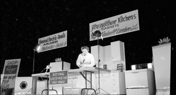 Ruth Hogan, a home economist with the National Livestock and Meat Board prepares a dish during a demonstration at the three-day "Rhapsody of Recipes" Cooking School at the Orpheum Theater. She is standing on stage in front of a kitchen setting with an array of appliances, cupboards, and advertisements for Kroger, General Electric, Madison Gas and Electric, Youngstown Kitchens and Cabinets, and Findorff Lumber and Supply.
