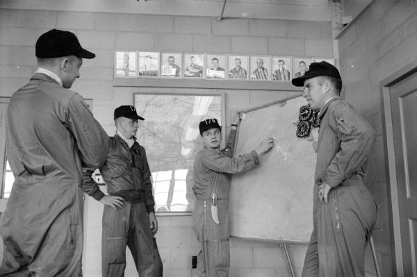 Air Guard Lt. Dan Torphy preparing for a training flight from Truax Field by attending a briefing for pilots by Capt. William Korbe of the 176th squadron operation officer. Dressed in their flight suits and baseball caps, they are (left to right): Dan Torphy, Lt. Phil Brickson, Capt. Korbe, and Lt. William Foy.