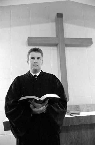 Reverend James Michael, holding a Bible and standing in front of a large crucifix, in the chancel of Our Redeemer Lutheran Church located at 5201 Old Middleton Road.