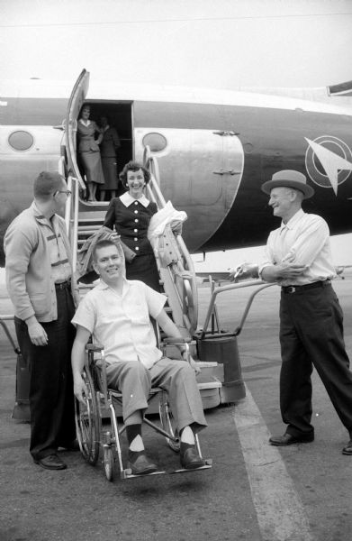 Robert Jensen, 24 of McFarland, is sitting in a wheelchair after exiting an airplane following a flight from Ann Arbor, Michigan, where he was treated for the respiratory complication of infantile paralysis brought on by polio. He is accompanied by Mrs. Merwyn Jachartt, a nurse from Ann Arbor. Greeting him are his father, Nels Jensen (right), and a friend Ras Kalnes.  