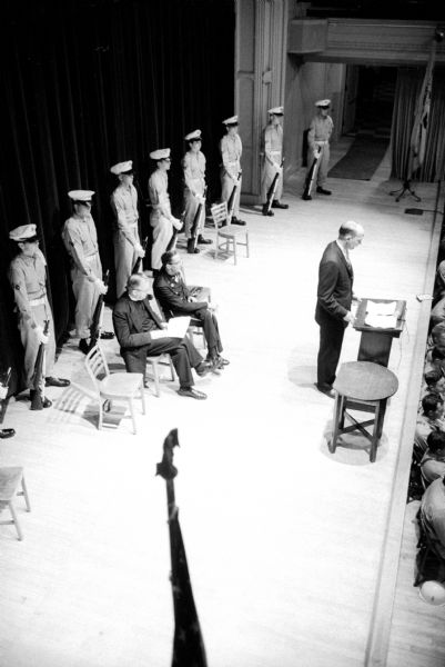 Elevated view of a group of people on a stage. Standing at a podium is Mayor Ivan Nestingen, speaking at the Memorial Day ceremony held at Central High School because of rain. Sitting left to right: Reverend Carl W. Stromberg, pastor of First Methodist Church, and Charles Suther, general chairman of the event. In the rear is standing the Truax Field Air Force honor guard.