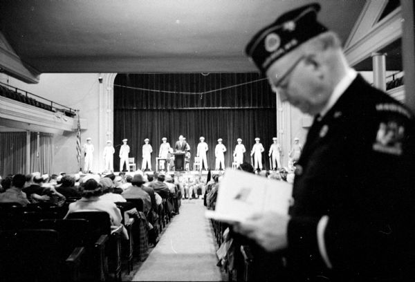 In the foreground a Veteran of Foreign Wars is looking at a Memorial Day program, held at Central High School because of rain. In the background is the audience, and on stage is Mayor Ivan Nestingen, flanked by an Air Force honor guard, delivering the ceremony's address.