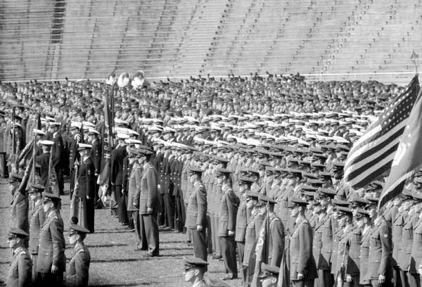 University of Wisconsin ROTC men stand at attention during a review at Camp Randall stadium. Empty bleachers fill the background and two flags are flying in the foreground. 