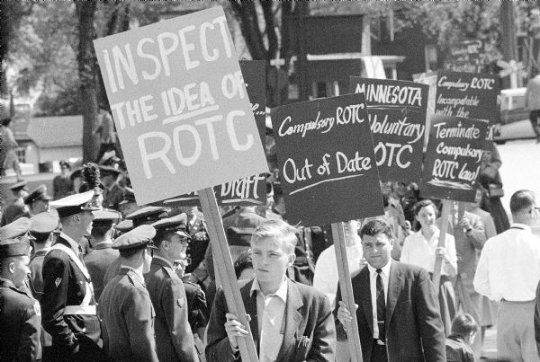 Anti-military protestors demonstrating outside U.W. Camp Randall stadium during a review of ROTC cadets. Posters challenge compulsory service in the state and reference the Wisconsin Idea.