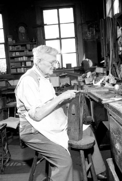 Adolph Frederick Schmidt sitting at his bench, working on a leather harness in his shop at 304 East Main Street. He started his career as a harness maker in 1895 when he was 19 years old. He will close his shop in three months.