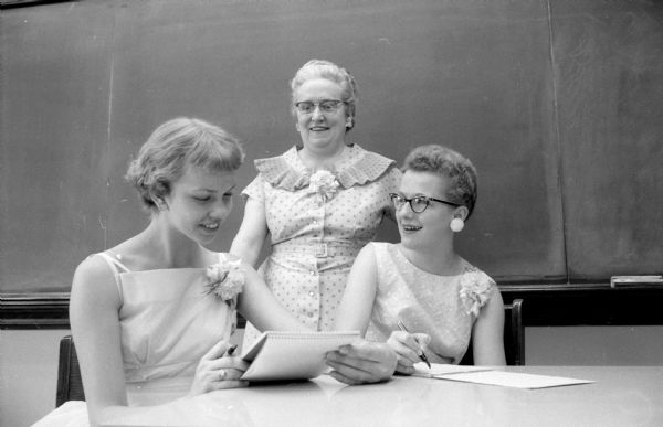 Madison East High School teacher in the school's business education department, Mary Heyer (standing by the chalkboard), awards gold pins to Barbara Lien (left) and Virginia Domini for their shorthand writing skills. They qualified for the pins by being able to take dictation at the rate of 160 words per minute for five minutes. The three have corsages pinned to their dresses.