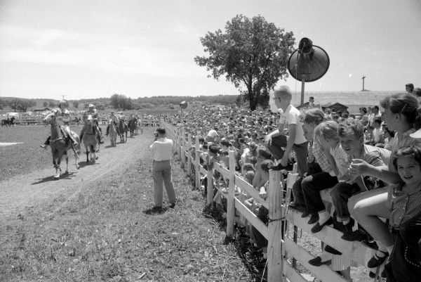 A packed crowd of children are watching from behind a white fence towards an arena of horseback riders. A large speaker is on a pole for announcements.

Approximately 3,500 children, ages 3-13, and parents attend the annual Golden G. Ranchers' Club party held at the Bowman Farm Dairy on Fish Hatchery Road. Six acts entertained at the miniature rodeo. Ben Berger and John Schermerhorn were co-masters of ceremonies.