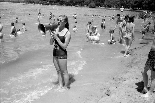 Lifeguard Nancy Thomsen (5524 Lake Mendota Drive) warns a youngster not to go into deep water at the Tenney Park beach on the official opening day for the summer of the Madison swimming beaches. Swimmers crowd the sandy Mendota lakeshore behind her. 