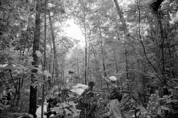 Sauk County Deputy Melvin Albers points out the direction that the light plane took as it crashed through a forest near Baraboo, taking the lives of Louis A. Gardner, president of the Gardner Baking Co., and his mother, Mrs. Louis Gardner. He is standing next to the wreckage of the fuselage and a crumpled wing surrounded by trees.