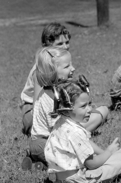 Three girls watching the neighbor kids at play at a city playground.  A girl in the foreground has pins holding her hair and the caption calls her a "Pin-Up."