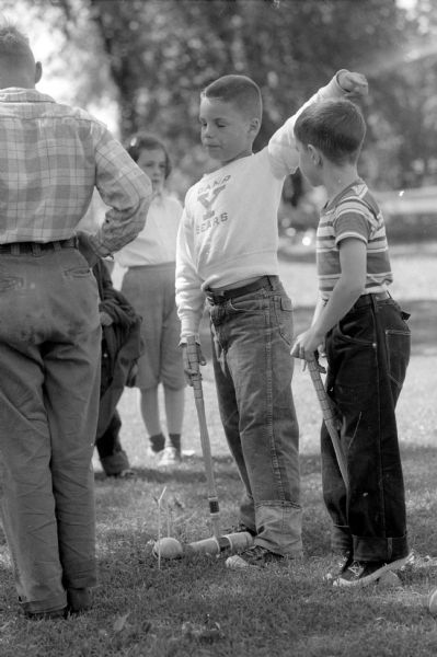 Children playing croquet at a city playground. The boy is pointing to where he is aiming to hit his ball with the mallet. The caption reads: "I'm going that-away!"