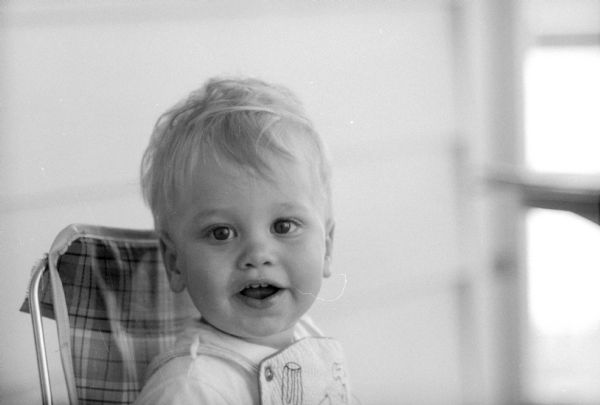Portrait of the baby son of <i>Wisconsin State Journal</i> photographer, Ed Stein. Jimmy Stein was born on June 8, 1956. He is sitting in a high chair and wearing overalls. 