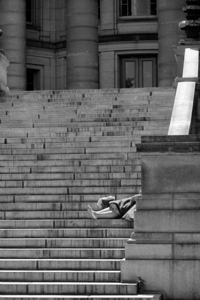 A partially hidden woman lounges in the sun on a flight of exterior steps of the Wisconsin State Capitol building. Only her legs and skirt are visible.