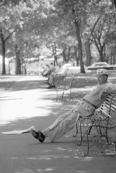 Man wearing a brimmed hat slumped alone on a metal park bench in the shade on South Pickney Street on Capitol Square. He has a cigarette or cigar between his fingers and a section of newspaper rests near his feet. Others are seen in the distance also sitting in the shade under trees.